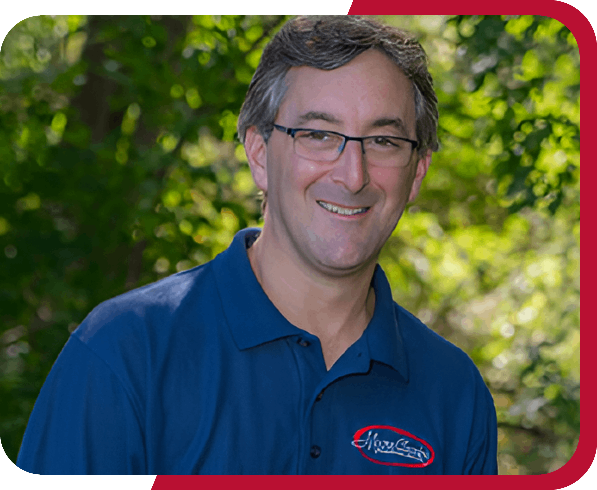 A man with glasses smiles, wearing a blue polo shirt with a logo. He stands outdoors with trees in the background.