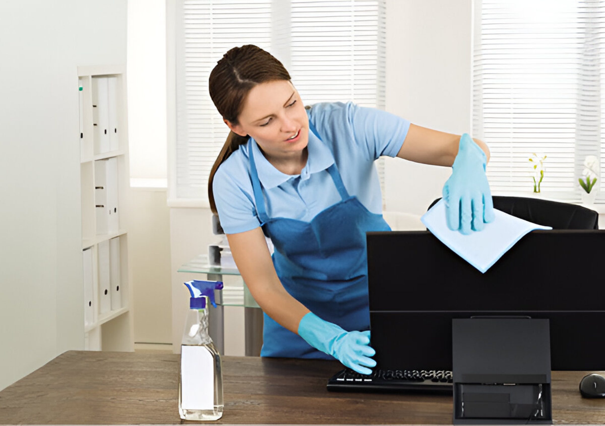 A woman in gloves cleans a computer monitor with a cloth in an office setting. A spray bottle is on the desk.