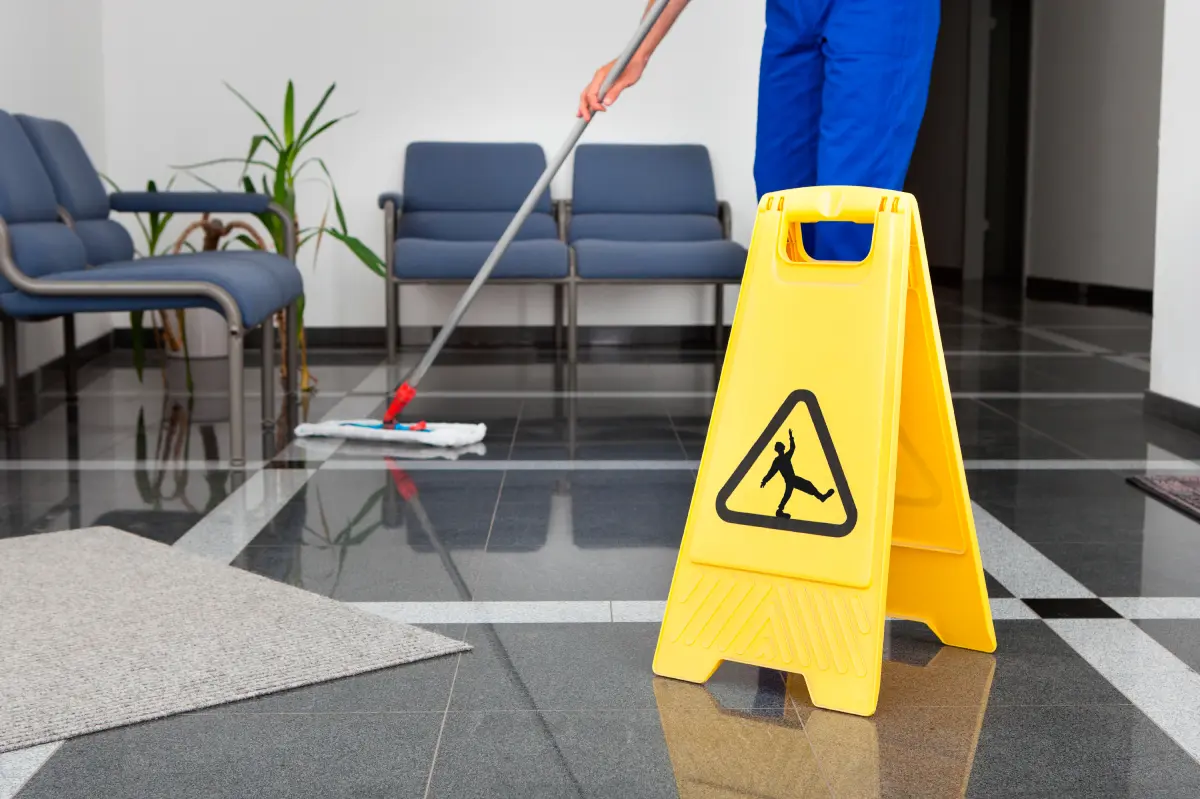 Person mopping the floor in a lobby area with a yellow caution sign in the foreground and gray chairs in the background.